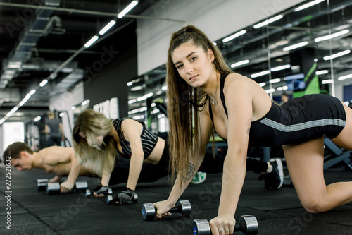 The athlete woman wrung out on dumbbells and looks into the camera. Young man and two woman training together with barbells in the gym photo