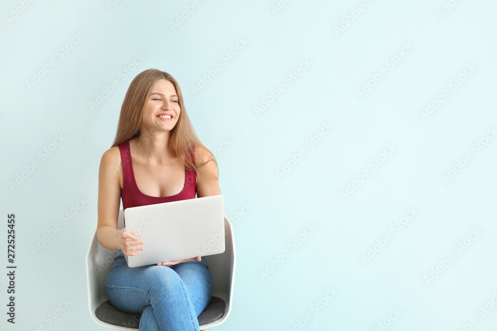 Beautiful young woman with laptop sitting on chair against color background Beautiful young woman with laptop sitting on chair against color background
