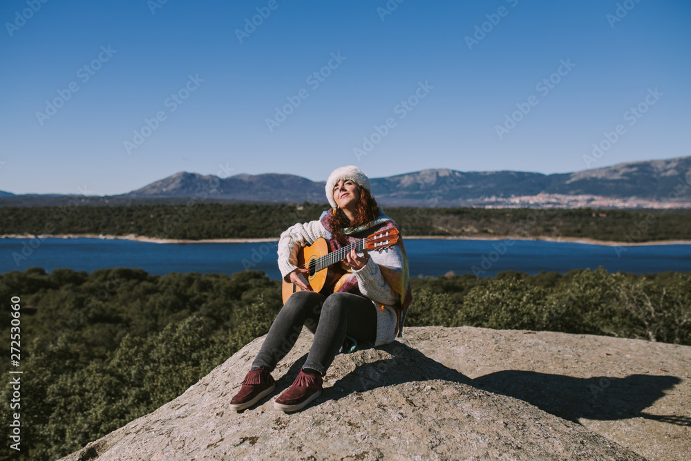 A beautiful young woman is sitting on the stone while playing the guitar. Behind her there is a beautiful landscape of mountains and a big lake. She is alone in the middle of the nature.