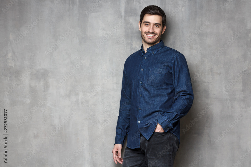 European male leaning on gray wall in denim shirt, looking confidently at camera