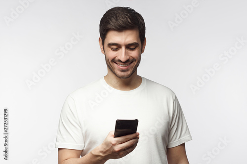 Closeup of Caucasian man holding phone in one hand and smiling happily while looking at screen, isolated on grey background © Damir Khabirov
