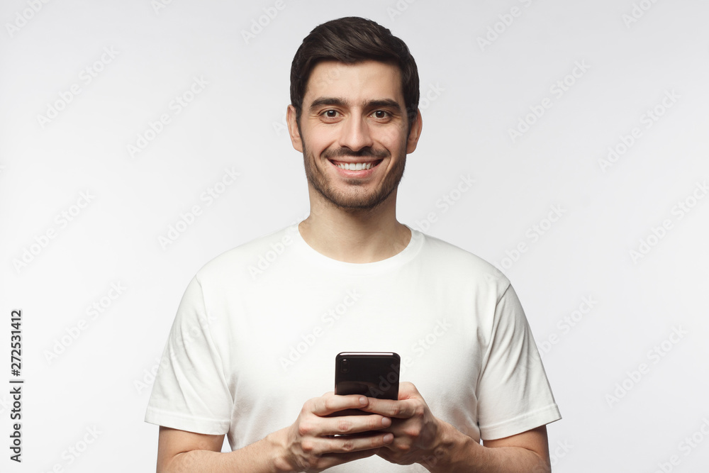 Young handsome man holding smartphone and looking at camera with smile while standing isolated on gray background Young handsome man holding smartphone and looking at camera with smile while standing isolated on gray background