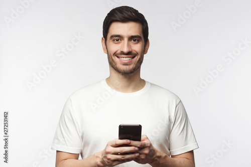 Young handsome man holding smartphone and looking at camera with smile while standing isolated on gray background © Damir Khabirov