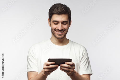 Portrait of handsome young man in white t-shirt, holding smartphone, playing video game, isolated on gray background