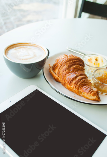 Tablet  fresh crispy croissant with jam  butter and cappuccino with beautiful latte art on grey table in a cafe. Morning routine.