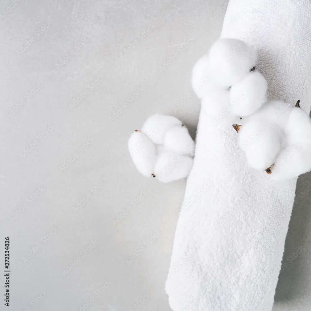 White fluffy towel twisted into roll with flowers of natural cotton on gray background with copy space. Spa concept. Flat lay.