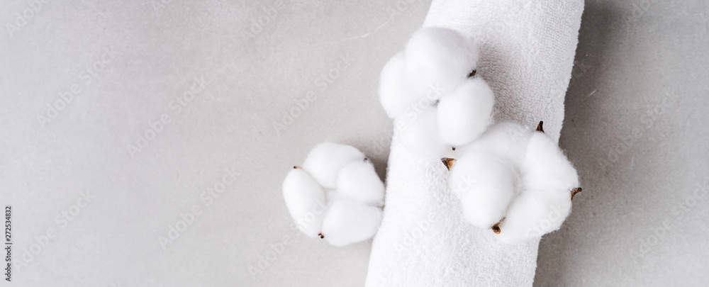 White fluffy towel twisted into roll with flowers of natural cotton on gray background with copy space. Spa concept. Flat lay. long wide banner White fluffy towel twisted into roll with flowers of natural cotton on gray background with copy space. Spa concept. Flat lay. long wide banner