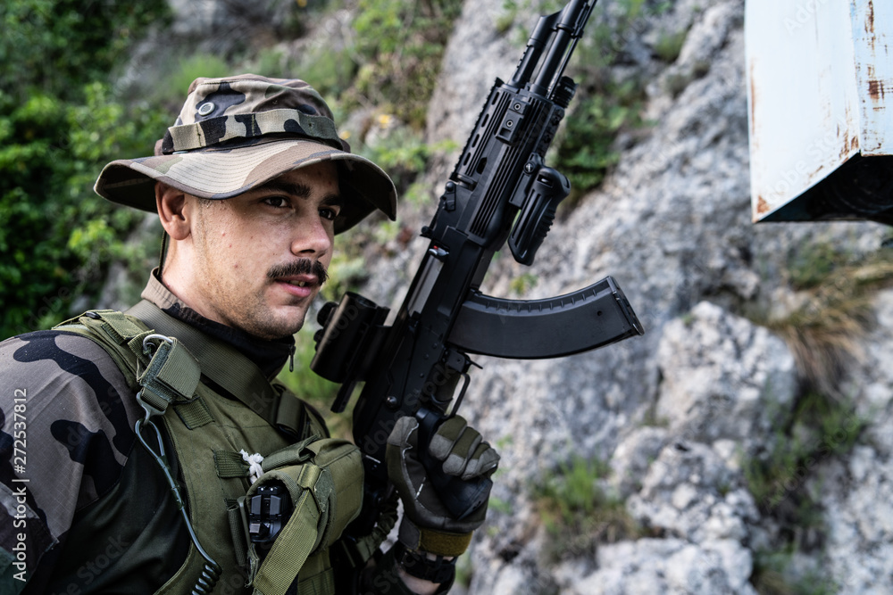 Young soldier with mustaches holding automatic machine gun in front of the mountain rocks