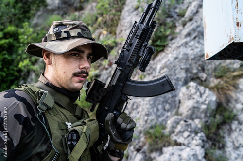 Young soldier with mustaches holding automatic machine gun in front of the mountain rocks