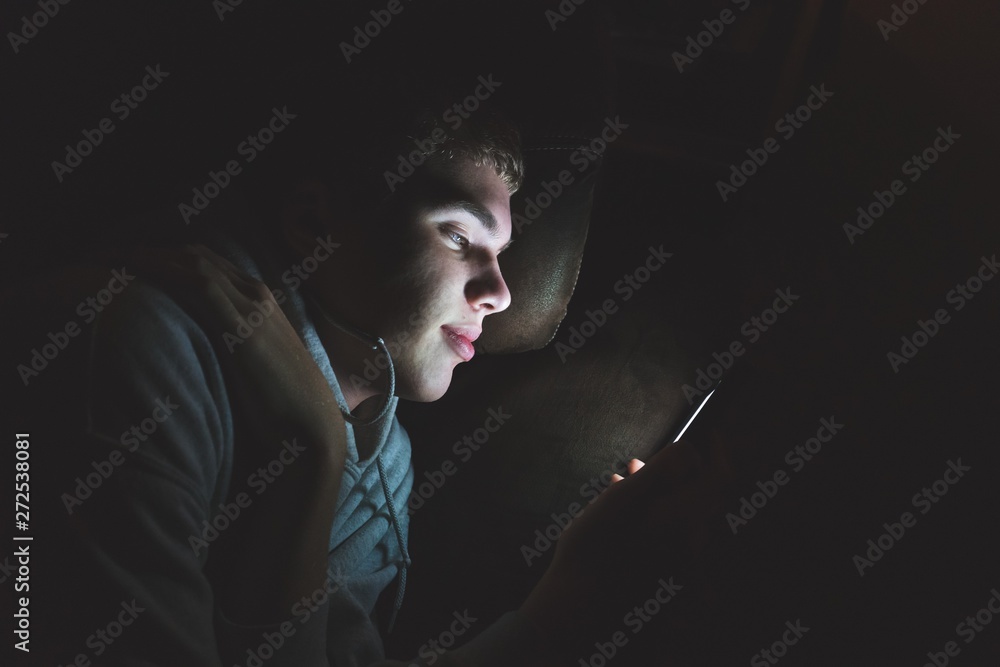 Obraz premium Teenager lying down on a couch in the dark. The light from the screen of his smartphone is illuminating his face.