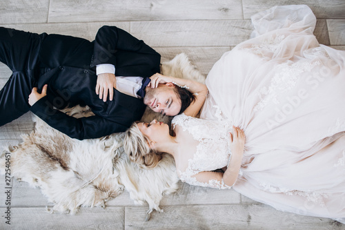 Bride and groom in wedding clothes are lying on the floor and smiling.