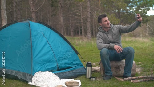 A young guy sits on a big log near the blue tent and takes a selfie on the phone. Background - pine forest.
