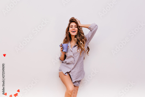 Sensual girl with light-brown wavy hair enjoying weekend morning. Spectacular female model in striped shirt holding blue mug with coffee.