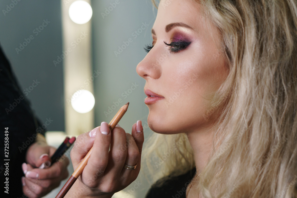 Obraz premium Make-up artist working in a make-up studio, applying makeup on the face of female clients. Makeup artist applies lipstick with a pencil. Evening make-up.