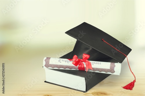 Graduation hat with book and diploma on wooden desk