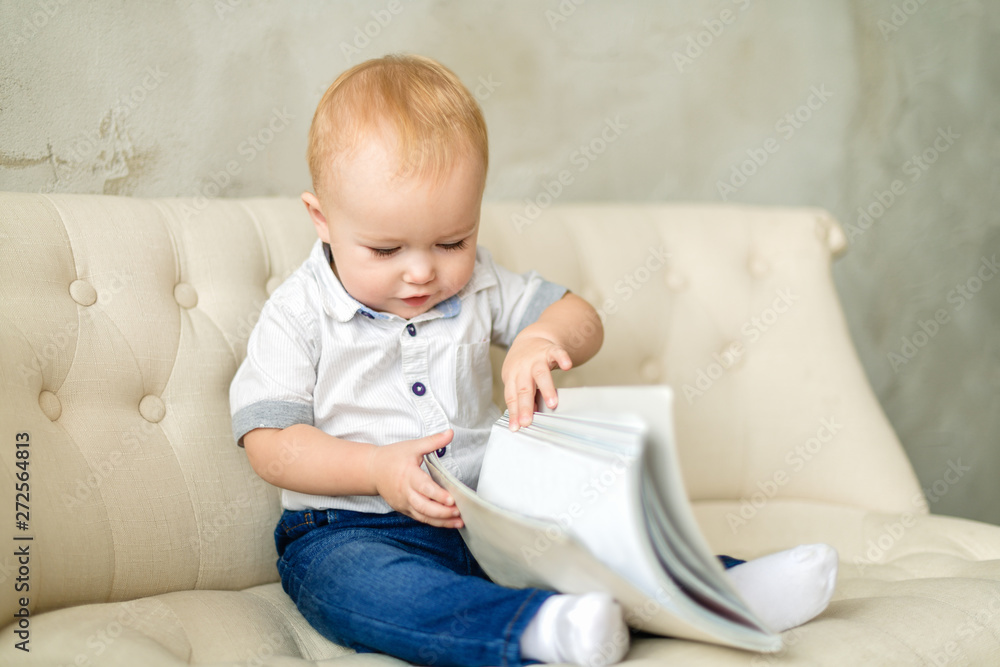 Happy chubby blond boy sitting on a light sofa and looking at a book. A red-haired boy in blue jeans and a white shirt is looking at a closed book. Horizontal photography Happy chubby blond boy sitting on a light sofa and looking at a book. A red-haired boy in blue jeans and a white shirt is looking at a closed book. Horizontal photography