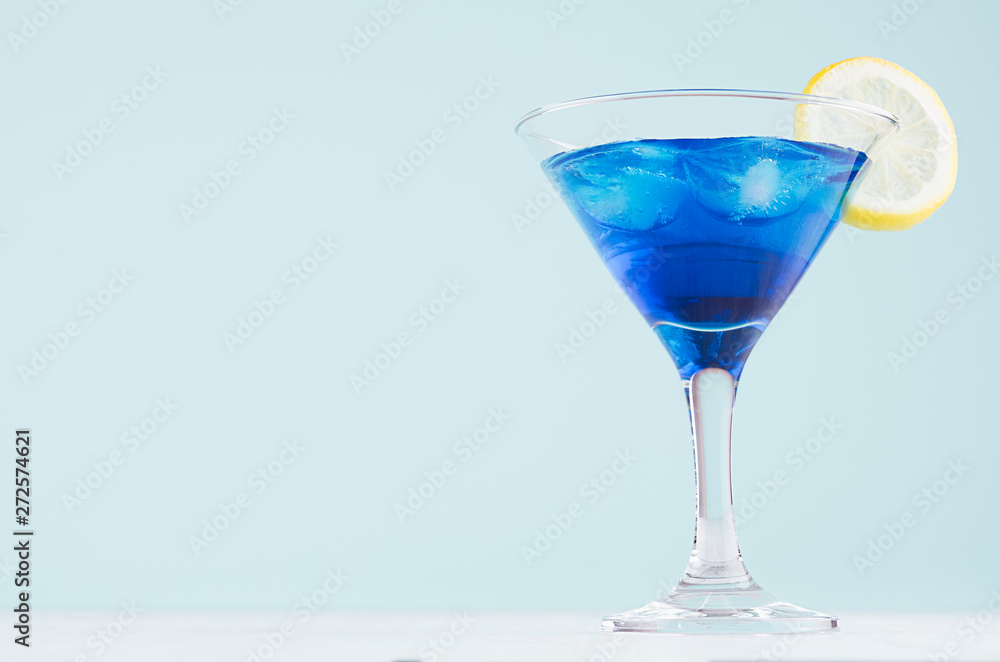 Blue curacao cocktail with ice cubes, lemon slice in luxury martini glass on white wood board and pastel mint color wall. Blue curacao cocktail with ice cubes, lemon slice in luxury martini glass on white wood board and pastel mint color wall.