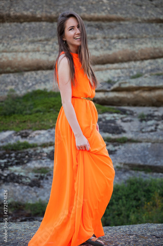 Young woman in orange long dress outdoor. Rocks and grass background. © Yuriy Pankratov