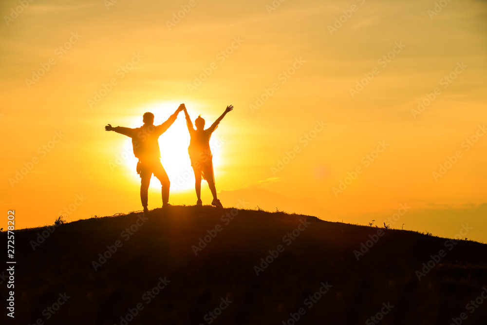 The couple stands holding hands on the top of the mountain with beautiful sunsets.happy man and woman tourist at hill,Happy couple with raised arms stands