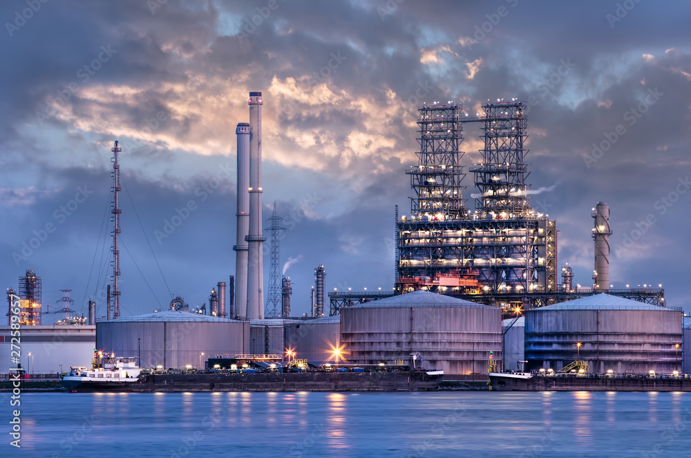 Petrochemical industry next to a river with a dramatic cloudy sky at twilight, Port of Antwerp, Belgium.