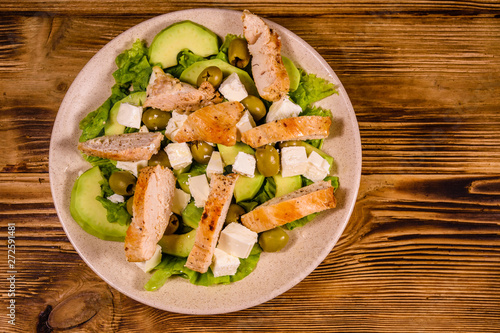 Fresh salad with chicken meat, feta cheese, avocado, green olives and lettuce leaves in ceramic plate on wooden table. Top view