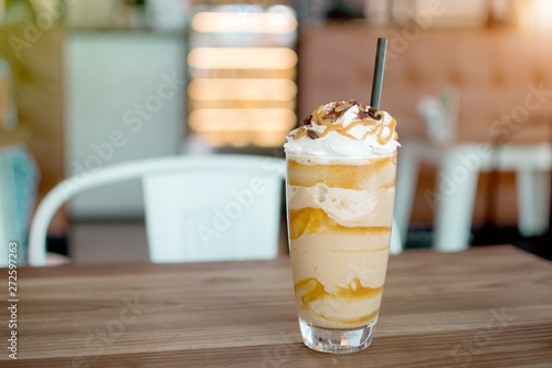 Iced frappe with whipped cream and caramel syrup, with straws and grains of coffee in a tall glass on the wooden table, cold coffee drink