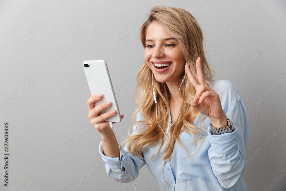 Young pretty blonde business woman posing isolated grey wall background using mobile phone listening music with earphones talking take a selfie. Young pretty blonde business woman posing isolated grey wall background using mobile phone listening music with earphones talking take a selfie.