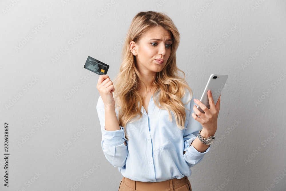 Sad young pretty blonde business woman posing isolated grey wall background holding credit card using mobile phone. Sad young pretty blonde business woman posing isolated grey wall background holding credit card using mobile phone.