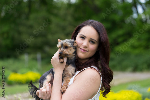Horizontal medium shot of cute smiling brunette young woman in white top holding small Yorkshire Terrier dog leaning against her