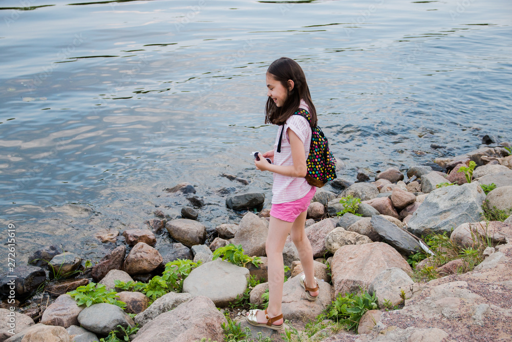 A girl with a backpack stands on the shore of a calm pond, next to large oval dark stones and germinating grass