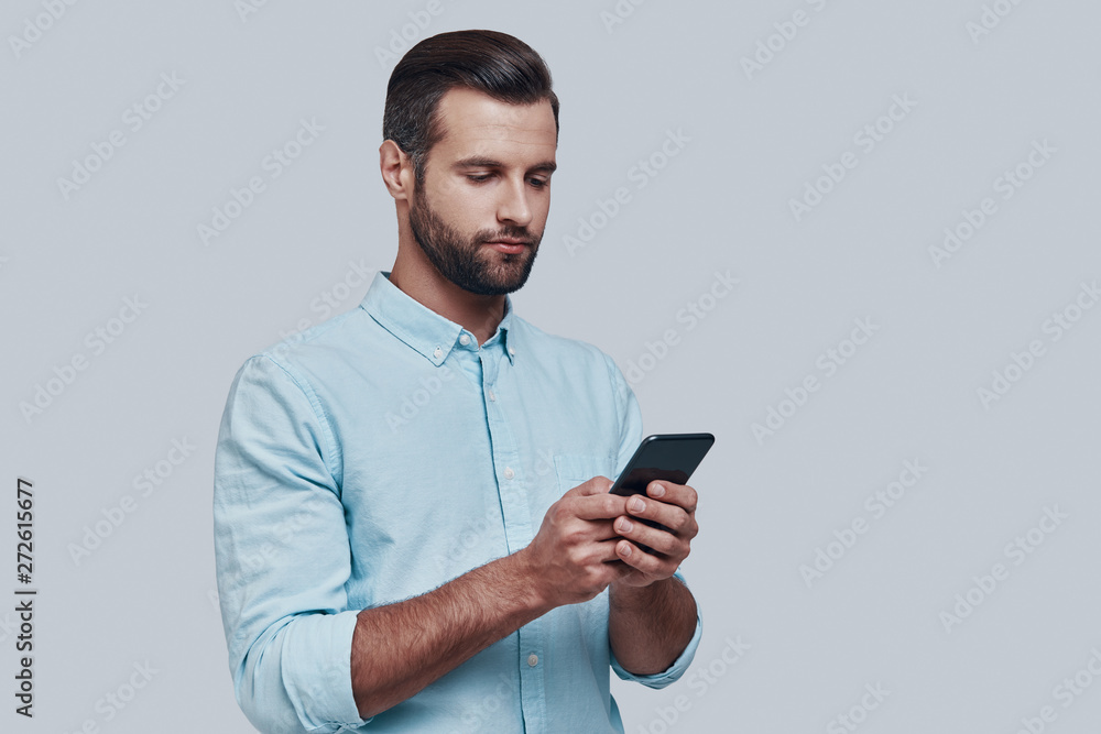 Checking messages. Handsome young man using smart phone while standing against grey background Checking messages. Handsome young man using smart phone while standing against grey background