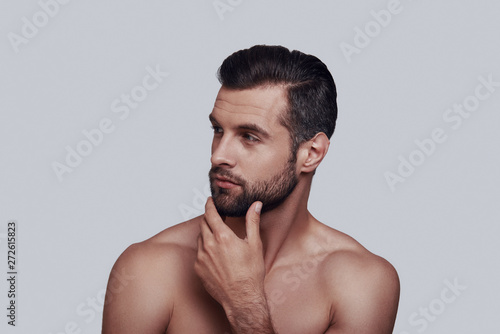 Time to shave. Handsome young man touching his beard while standing against grey background