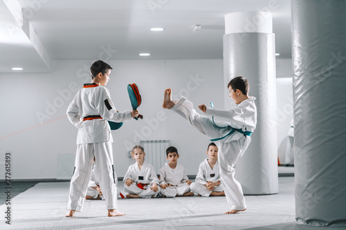 Two young Caucasian boys in doboks having taekwondo training at gym. One girl kicking while other one holding kick target. In background their friend sitting with legs crossed and watching them.