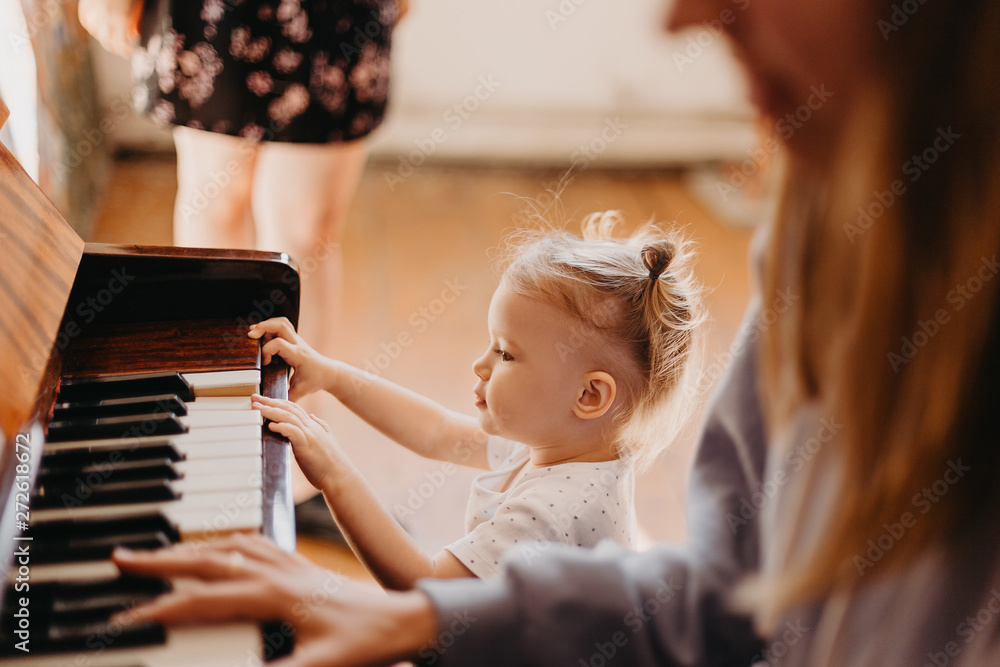 Mother teaches a little girl to play the piano. They play and sing songs. Its fun, Cute happy child girl playing piano in a light room. Selective focus, noise effect - obrazy, fototapety, plakaty 