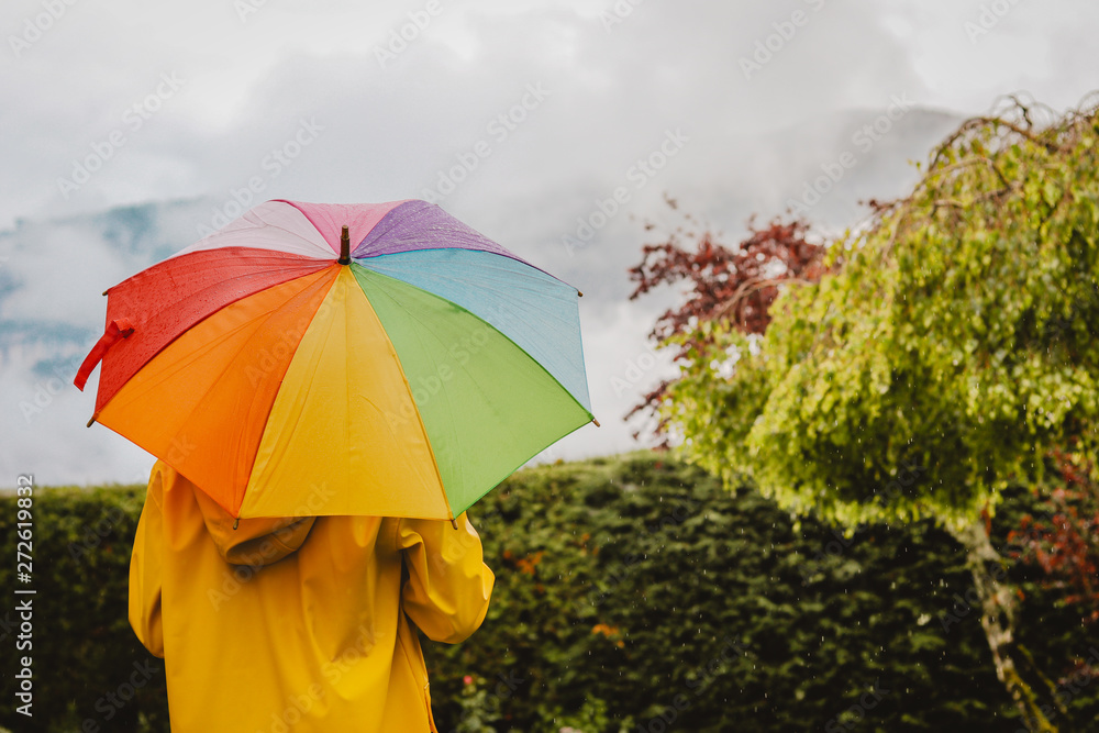 Back view of girl in yellow raincoat with colorful rainbow umbrella in park outdoors under rain looking at cloudy sky and mountain. Rainy summer weather concept