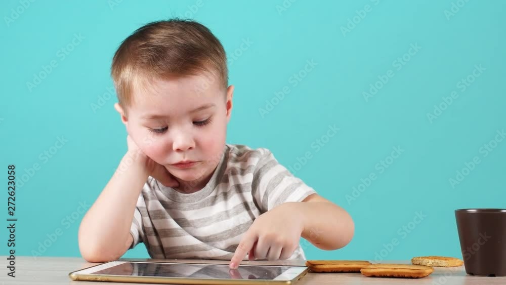 Close up serious face cute little child uses a tablet PC sitting at table, isolated on blue Close up serious face cute little child uses a tablet PC sitting at table, isolated on blue