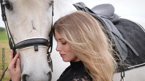 closeup of a beautiful blonde girl with black dress strolls in the meadow with a white horse