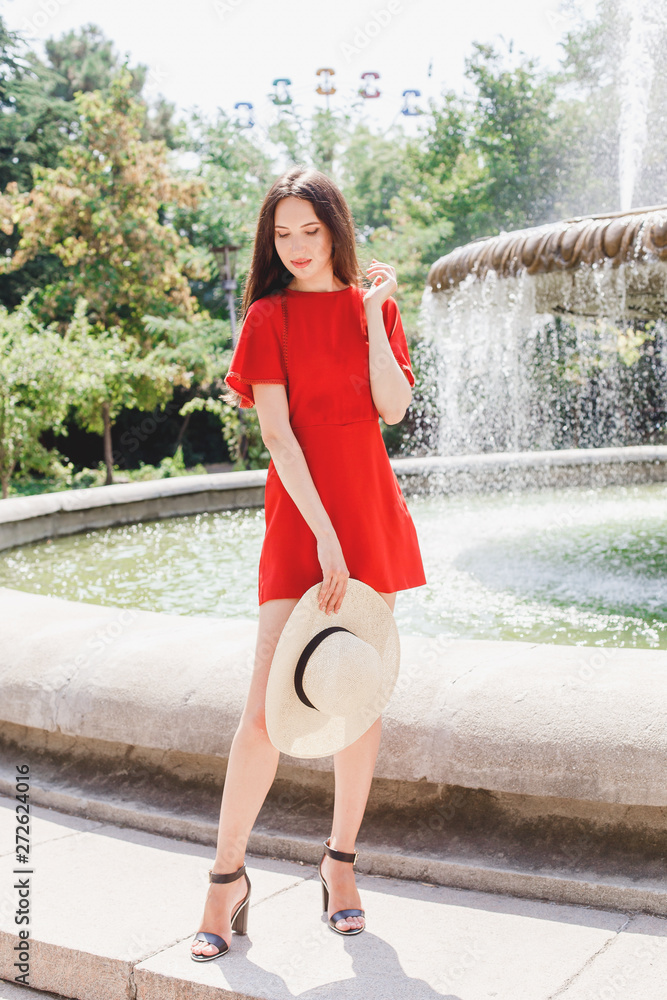 Young stylish beautiful brunette woman, wearing red dress, black high heel  sandals and straw hat, standing outdoors near fountain in the city. Trendy  casual summer outfit. Street fashion. Stock Photo | Adobe