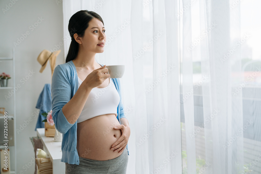 young healthy asian pregnant woman in sports bra and leggings standing by  window touching belly as