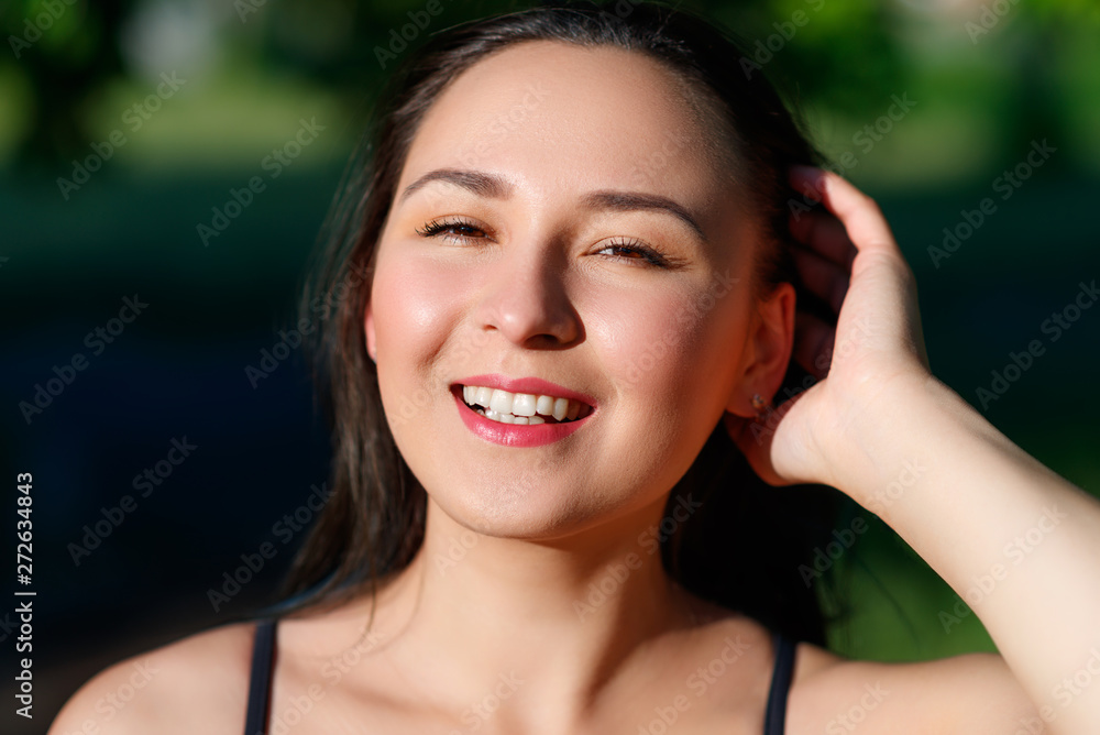 Fototapeta premium close up portrait of a young beautiful smiling happy brunette girl in outdoor Park on a Sunny summer day correcting hand loose hair