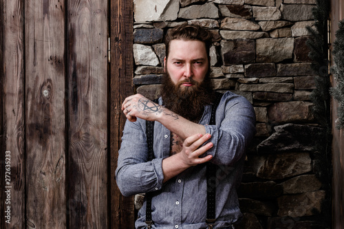 Brutal strong man with a beard and tattoos on his hands dressed in stylish casual clothes stands on the background of stone wall and wooden door