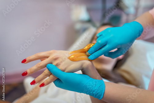 Close-up hands of cosmetologist in blue gloves applying golden paste for sugaring depilation on woman arm  hair removal beauty procedurein spa salon.