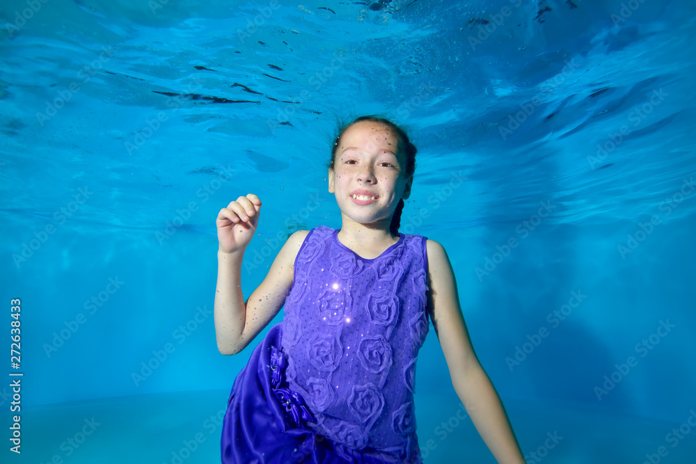 A little girl swims underwater in the pool. She smiles, looks at the camera with her eyes open. She's wearing a beautiful dress. Blue background. Portrait. Horizontal view