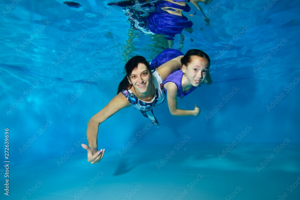 Mom and little daughter swim underwater in the pool. They hug, smile, look at the camera with their eyes open. They're wearing beautiful dresses. Portrait. Horizontal view