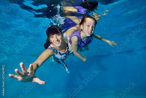 Mom and her little daughter are swimming underwater in the pool. They hug, smile, look at the camera with their eyes open. Portrait. Horizontal view