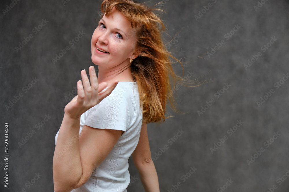 Fototapeta premium Studio portrait of a beautiful pretty woman age of 40 years on a gray background, smiling, showing different emotions, happy with life. In a white T-shirt with red hair. Directly opposite the camera.