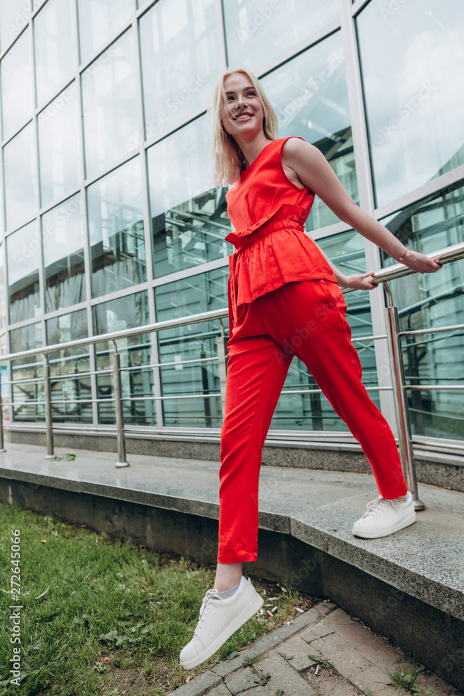 young smiling woman with light hair dressed in a bright red suit with pants and white sneakers on the background of a glass building, urban clothing style. Street photography