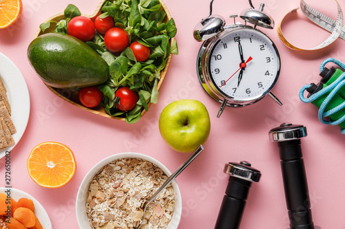 top view of fresh diet food, measuring tape, sport equipment and alarm clock on pink background with copy space