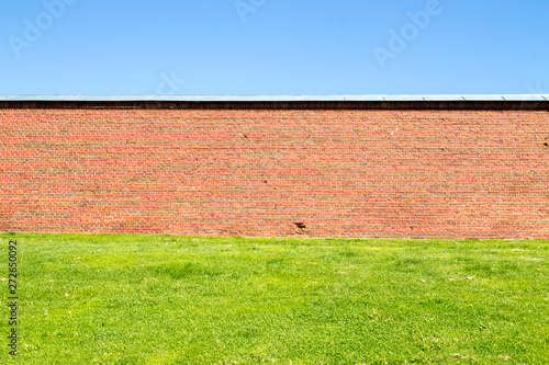 Old red brick wall behind a field of grass against clear blue sky background