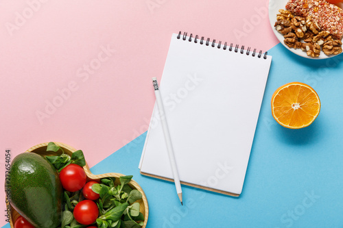 top view of empty notebook, vegetable salad, orange and nuts on pink and blue background